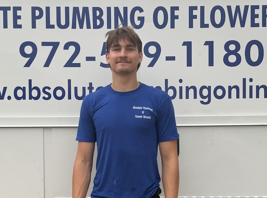 Young man in a blue t-shirt stands in front of a white sign advertising Absolute Plumbing of Flower Mound with a large phone number above.