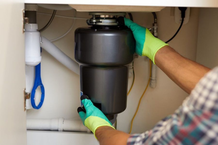 Gloved hands of a worker installing or repairing a garbage disposal under a kitchen sink.