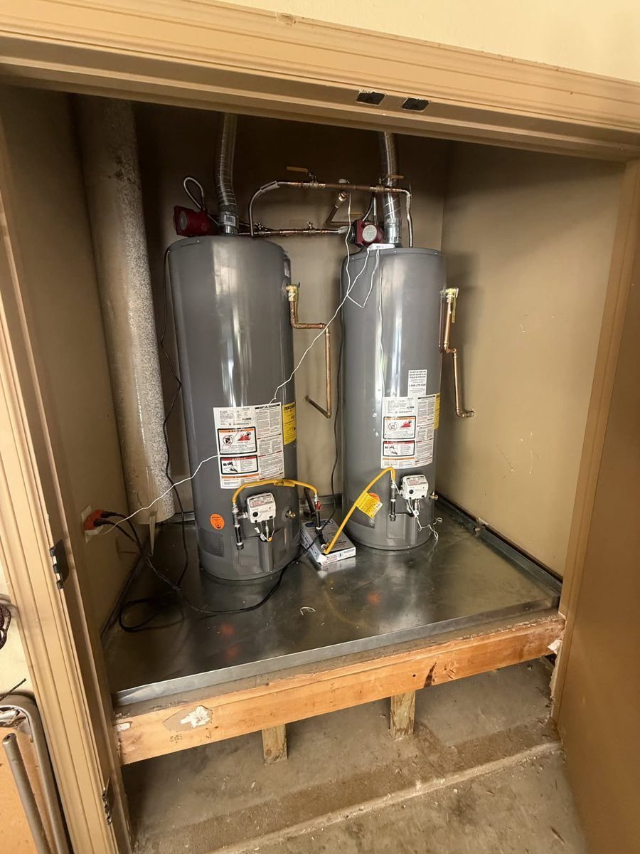 Two tall gray water heaters stand side by side in a small utility closet, with copper piping, gauges, and a vent pipe nearby on the left.
